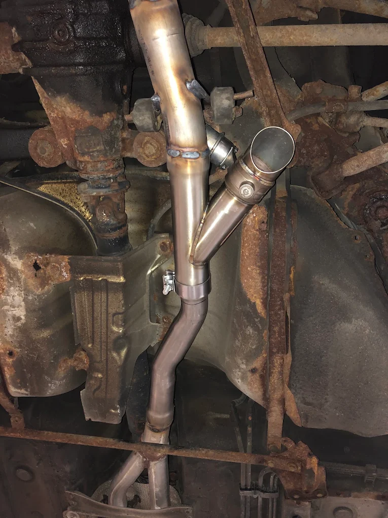Newly welded stainless exhaust Y-pipe fitted against the heavily rusted original underbody of a vehicle