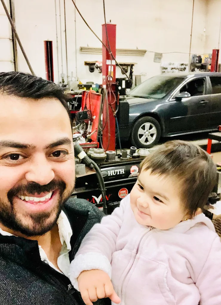 Pete, owner of Pete's Muffler & Automotive, holding his daughter in the Malcolm Rd shop with a vehicle on the hoist behind him